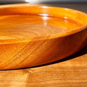 Puede incluir: Primer plano de un cuenco de madera para servir con un cálido tono marrón rojizo. El cuenco tiene una superficie lisa y pulida y un borde redondeado. La veta de la madera es visible, añadiendo textura. Se encuentra sobre una superficie de madera.