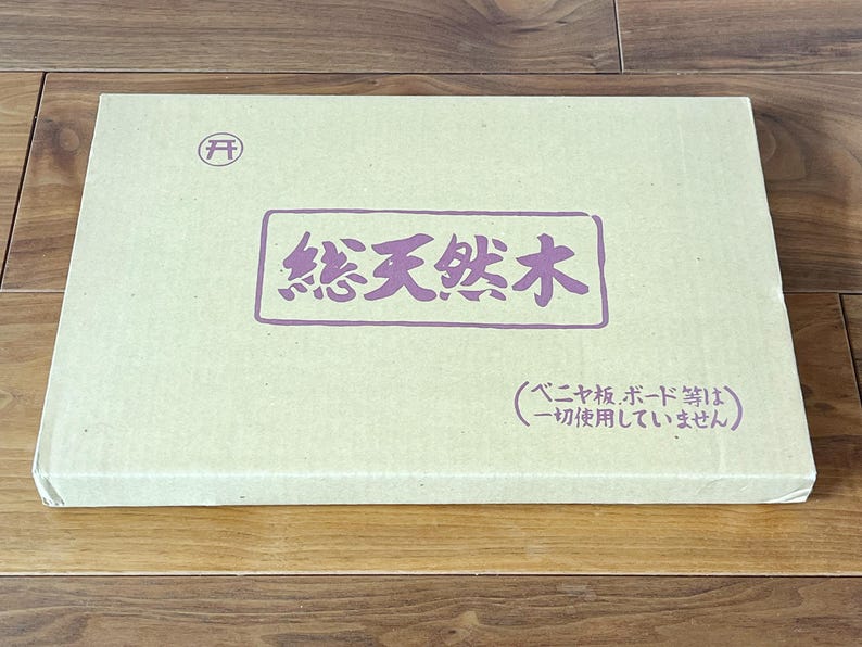 May include: A rectangular, light beige box with Japanese text in a purple rectangle. The text reads "Total Natural Wood". A small circular symbol is in the upper left corner. The box is on a wooden surface.