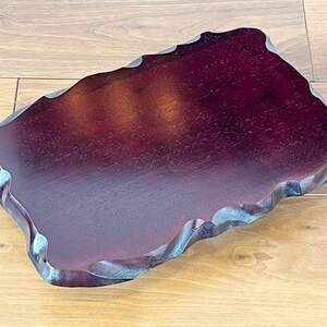 May include: A dark burgundy wooden tray with a distinctive, wavy edge. The tray has a smooth, polished surface and a natural, organic shape. The wood grain is visible, adding to its rustic charm. The tray is suitable for serving or display.
