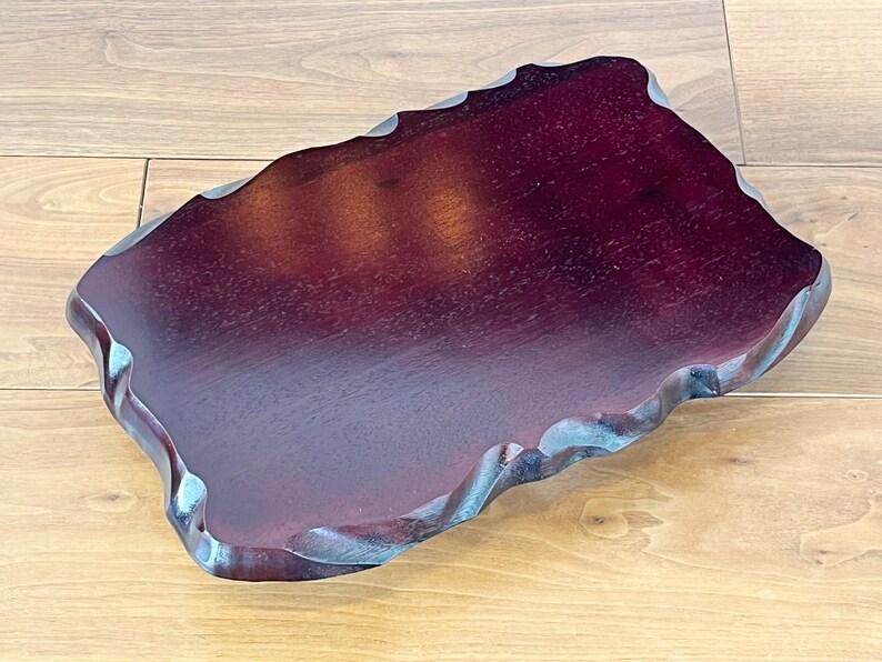May include: A dark burgundy wooden tray with a distinctive, wavy edge. The tray has a smooth, polished surface and a natural, organic shape. The wood grain is visible, adding to its rustic charm. The tray is suitable for serving or display.