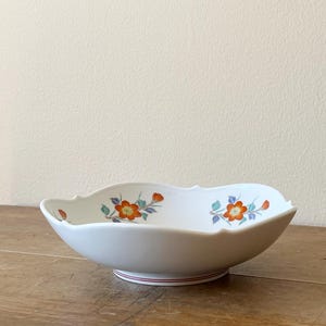 May include: A white ceramic bowl with a scalloped edge and a floral design featuring orange and blue flowers. The bowl is sitting on a wooden surface.