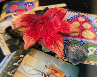 Puede incluir: Un plato de cerámica de hoja de arce rojo y naranja con un diseño en relieve. El plato está colocado sobre una pila de cartas de tarot. Las cartas presentan ilustraciones coloridas y texto, incluyendo "Tres de Copas" y "La Emperatriz".