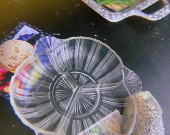 Puede incluir: Un plato de servir de vidrio transparente dividido con un diseño radial. El plato tiene tres secciones y bordes decorativos. La imagen también muestra una concha y una bandeja decorativa con alimentos.