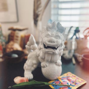 May include: White ceramic foo dog figurine with a snarling expression, standing on a round base. The figurine is on a black surface with a pink flower and a card with colorful graphics.