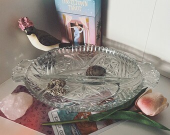 Puede incluir: Un plato de servir de vidrio con un patrón decorativo, un tulipán rosa, una carta de tarot con el texto "King of Cups", un cristal rosa, una pequeña figura de pájaro y una caja de cartas "Tinseltown Tarot".