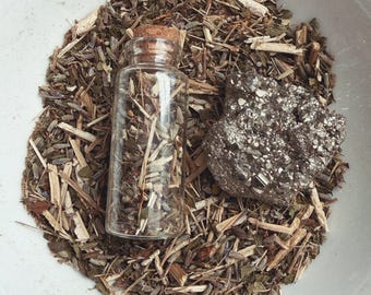 May include: A small glass bottle with a cork stopper filled with dried herbs, resting on a bed of similar herbs and a dark, textured stone. The arrangement is in a white bowl with a blue rim.