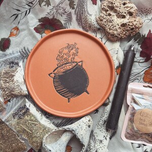 May include: An orange ceramic plate with a black cauldron design, surrounded by dried herbs in small bags, a black candle, a seashell, and a porous rock. The items are arranged on a floral patterned fabric, creating a mystical theme.