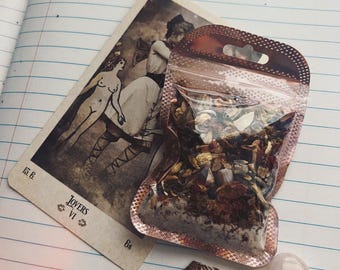 May include: A tarot card with the Lovers card image, a small bag of dried herbs, a seashell, and a heart-shaped crystal on a lined notebook.