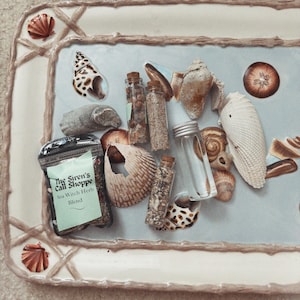 May include: A collection of seashells, small glass bottles, and a package of Sea Witch Herb Blend arranged on a rectangular tray with a light blue surface. The tray has a white border with seashell accents. The text "The Siren's Call Shoppe" is visible.