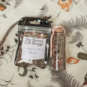 May include: A clear glass bottle and a clear plastic bag filled with dried herbs and flowers. The bag has a black top and a white label that reads "The Siren's Call Shoppe" and "Persephone Herb Blend". The bottle is sealed with pink wax.