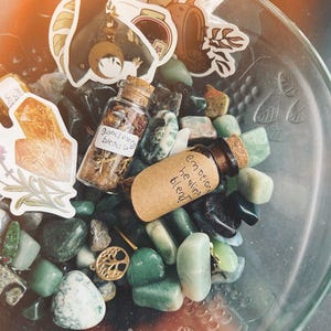 May include: A glass bowl filled with green and white stones, two small glass bottles with cork tops, and various stickers. The bottles are labeled "good energy blend" and "emotion healing blend".