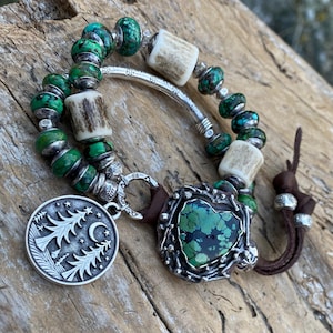 May include: Handmade bracelet featuring green turquoise beads, antler accents, and a silver charm with a tree and crescent moon design. The bracelet includes a heart-shaped turquoise stone and a brown leather cord.