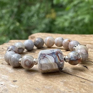May include: A beaded bracelet featuring a large, central, faceted cylindrical bead with swirling bands of white, grey, and brown, flanked by smaller, smooth, round, variegated agate beads in shades of grey, tan, and brown with silver spacers.