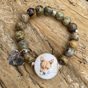 May include: A beaded bracelet featuring a round charm with a dog portrait. The bracelet includes various earth-toned, round beads and a silver heart charm. The dog charm is white with a brown and tan dog.