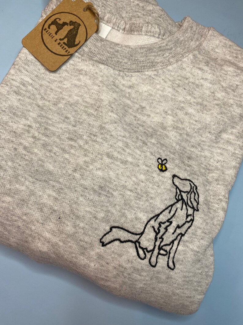 May include: A heather gray sweatshirt featuring an embroidered black dog looking up at a yellow and black bee. A brown tag with the text "Maisie & Murphy" is attached to the sweatshirt.