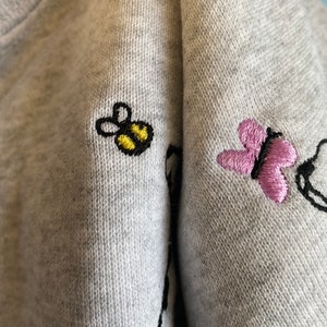 May include: Close-up of a gray sweatshirt with embroidered details. A yellow and black bee and a pink butterfly are embroidered on the fabric. The sweatshirt has a soft, textured appearance.