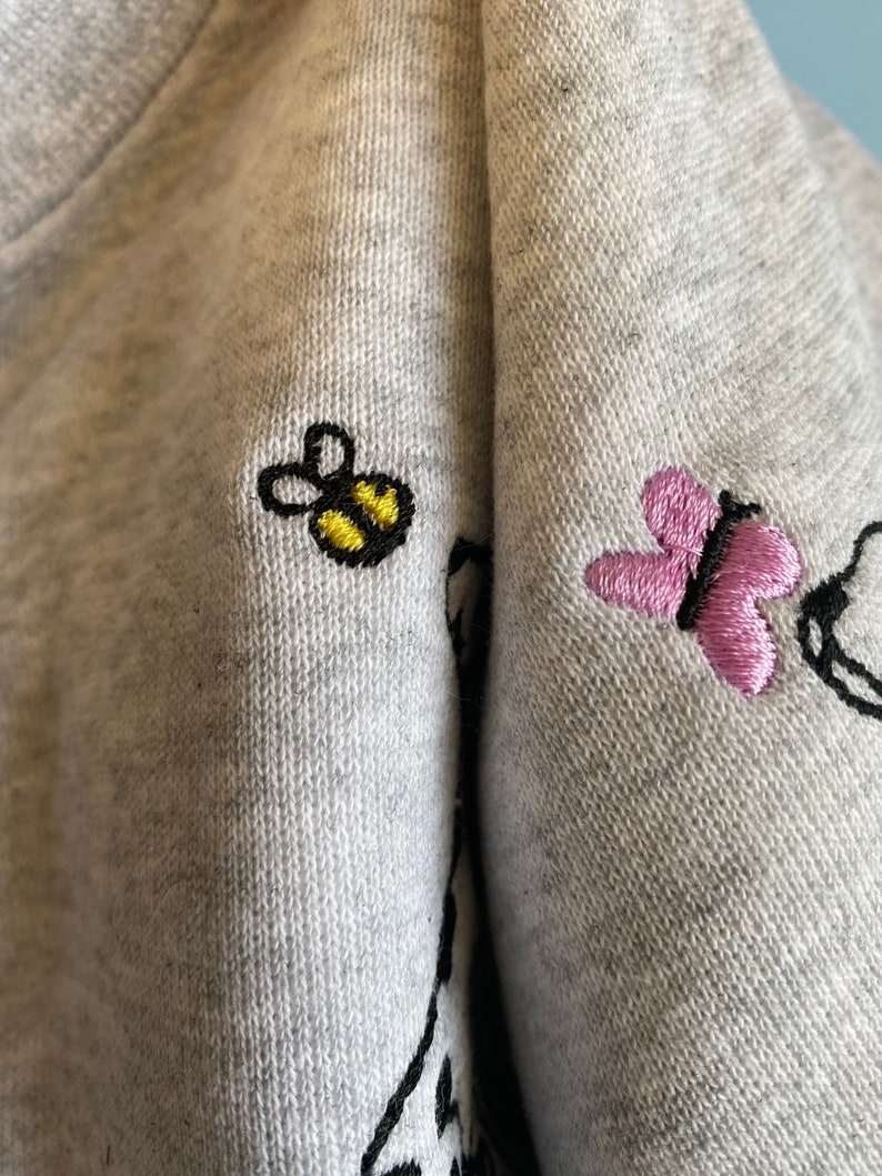 May include: Close-up of a gray sweatshirt with embroidered details. A yellow and black bee and a pink butterfly are embroidered on the fabric. The sweatshirt has a soft, textured appearance.