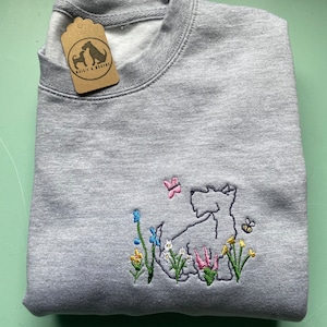 May include: Gray sweatshirt with an embroidered design of a white Scottish terrier dog in a field of flowers. The dog is surrounded by yellow, pink, blue, and green flowers. A butterfly and a bee are also embroidered on the sweatshirt. The sweatshirt has a tag that says "Westie & Buddy".