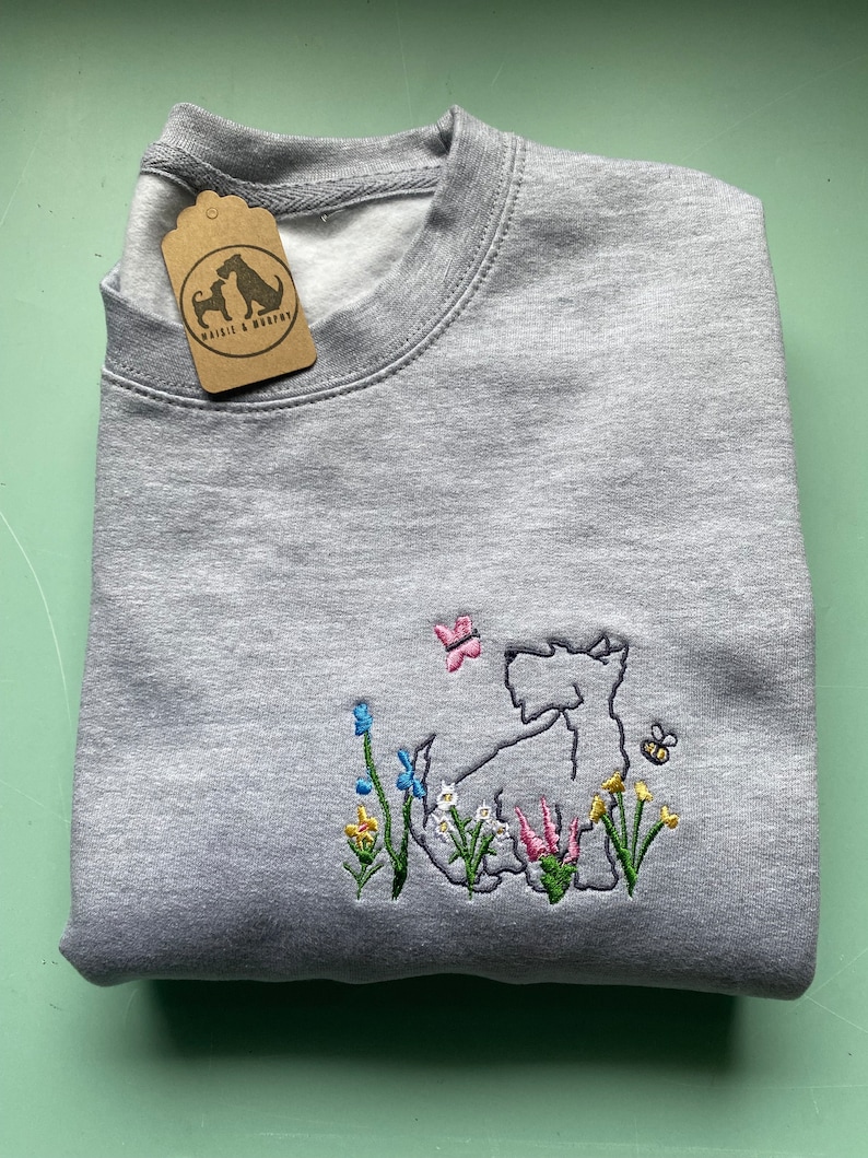 May include: Gray sweatshirt with an embroidered design of a white Scottish terrier dog in a field of flowers. The dog is surrounded by yellow, pink, blue, and green flowers. A butterfly and a bee are also embroidered on the sweatshirt. The sweatshirt has a tag that says "Westie & Buddy".