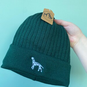 May include: A dark green ribbed knit beanie with a white embroidered Dalmatian dog design. The beanie has a folded brim and a hang tag with a logo. The hat is made of a soft, warm material.