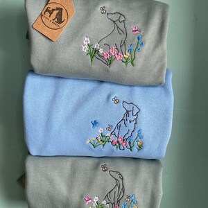 May include: Three gray and blue t-shirts with embroidered images of dogs looking up at bees. The shirts feature a floral design with pink, yellow, and blue flowers. The text "Whistle & Wag" is on a brown tag attached to the top shirt.