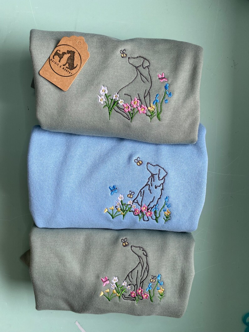 May include: Three gray and blue t-shirts with embroidered images of dogs looking up at bees. The shirts feature a floral design with pink, yellow, and blue flowers. The text "Whistle & Wag" is on a brown tag attached to the top shirt.