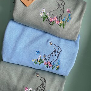 May include: Three sweatshirts with embroidered images of dogs looking up at bees. The sweatshirts are in shades of green and blue. The top sweatshirt has a brown tag with the text "Maisie & Maple".