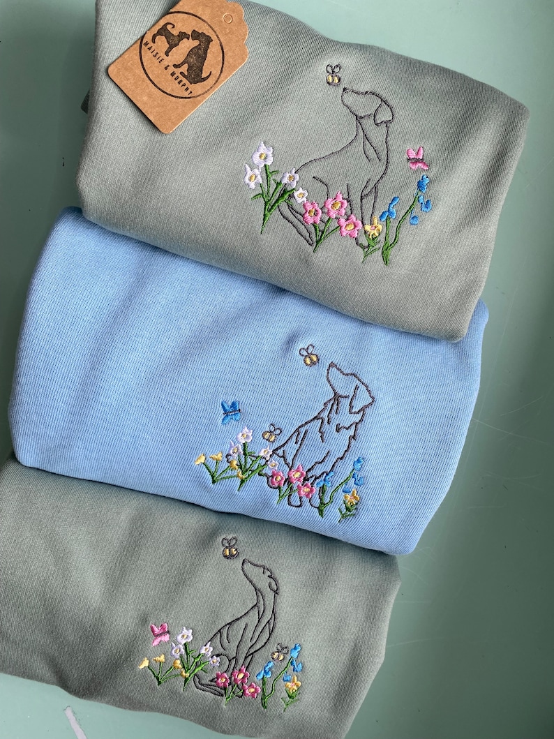 May include: Three sweatshirts with embroidered images of dogs looking up at bees. The sweatshirts are in shades of green and blue. The top sweatshirt has a brown tag with the text "Maisie & Maple".