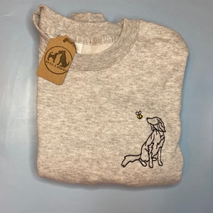May include: A light gray sweatshirt with a crew neck and a folded design. The sweatshirt features an embroidered dog looking at a bee. A brown tag with the text "Maisie & Murphy" is attached to the sweatshirt.