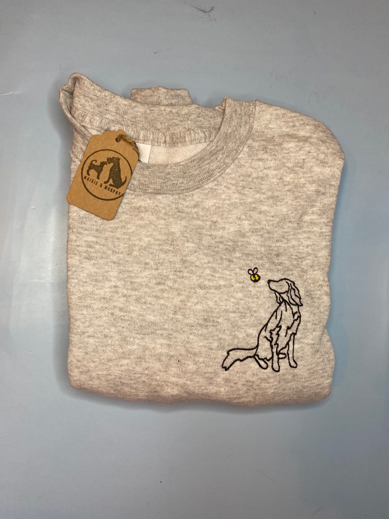 May include: A light gray sweatshirt with a crew neck and a folded design. The sweatshirt features an embroidered dog looking at a bee. A brown tag with the text "Maisie & Murphy" is attached to the sweatshirt.