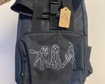 May include: Black backpack with a white embroidered design of three dogs. The backpack has a roll-top closure and a small brown tag with the text "Whistle & Growl".