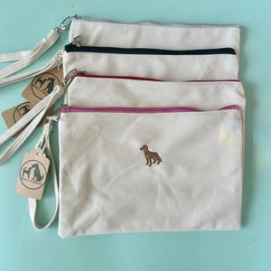 May include: Off-white canvas wristlet bags with colored zippers and a small embroidered dog. The bags have a wrist strap and a tag with a dog logo. The zippers are gray, black, red, and pink.