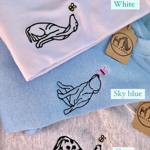 May include: White, sky blue, and grey sweatshirts with embroidered dog designs. Each sweatshirt features a different design: a dog reaching for a bumblebee or butterfly. The text "Colour options" and "choose whether you want a bumblebee or butterfly" are also visible.