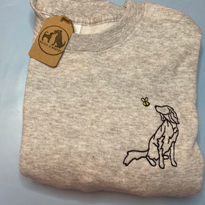 May include: A light gray sweatshirt with a crew neck and a folded appearance. The sweatshirt features an embroidered design of a dog looking up at a bee. A brown tag with the text "Waisle & Murphy" hangs from the sweatshirt.