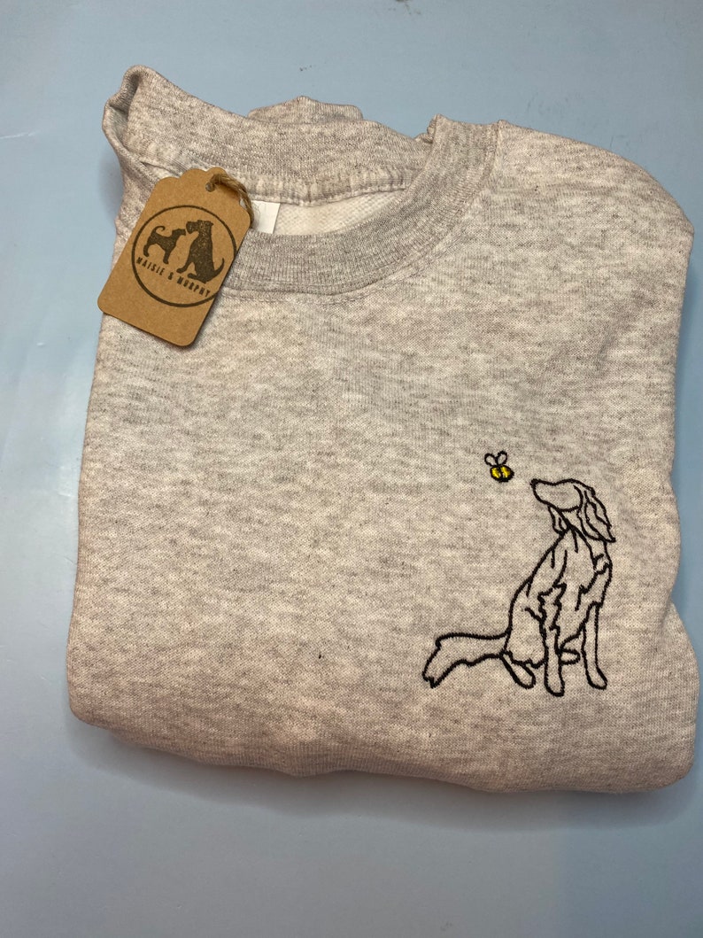 May include: A light gray sweatshirt with a crew neck and a folded appearance. The sweatshirt features an embroidered design of a dog looking up at a bee. A brown tag with the text "Waisle & Murphy" hangs from the sweatshirt.