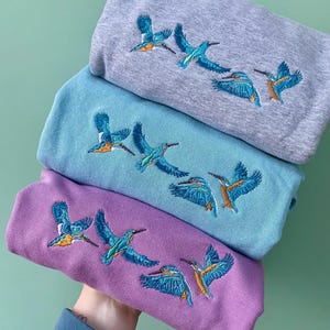 May include: Three sweatshirts with embroidered kingfisher birds. The sweatshirts are in shades of grey, blue, and purple.