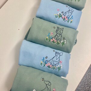 May include: Five sweatshirts stacked on top of each other. The sweatshirts are a light blue and a sage green color. Each sweatshirt has an embroidered design of a dog sitting in a field of flowers.
