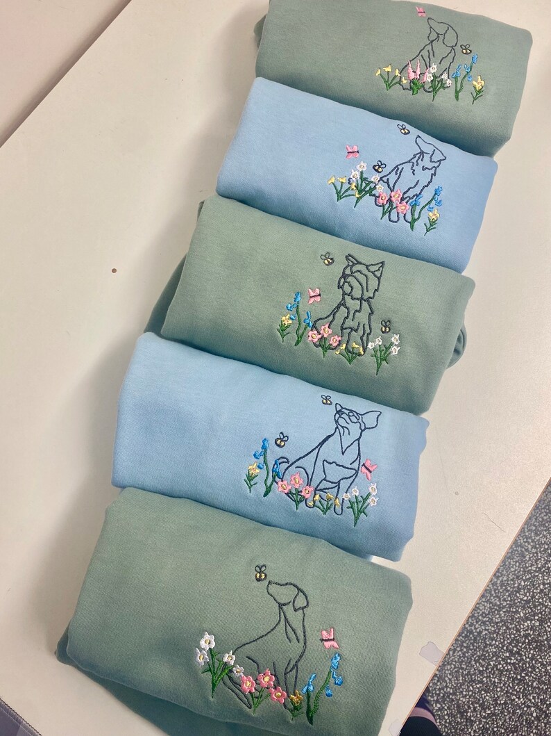 May include: Five sweatshirts stacked on top of each other. The sweatshirts are a light blue and a sage green color. Each sweatshirt has an embroidered design of a dog sitting in a field of flowers.