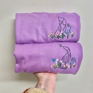 May include: Two lavender sweatshirts with embroidered images of a dog looking up at a butterfly and flowers. The dog is outlined in black and the flowers are in shades of pink, blue, yellow, and green.