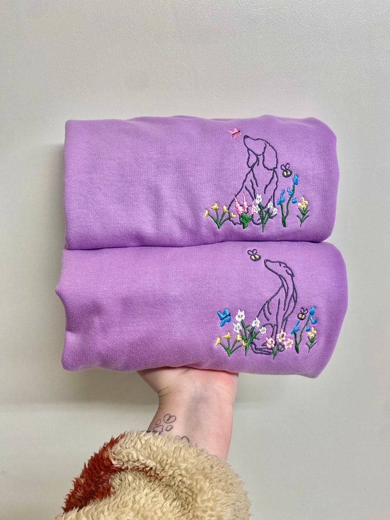 May include: Two lavender sweatshirts with embroidered images of a dog looking up at a butterfly and flowers. The dog is outlined in black and the flowers are in shades of pink, blue, yellow, and green.