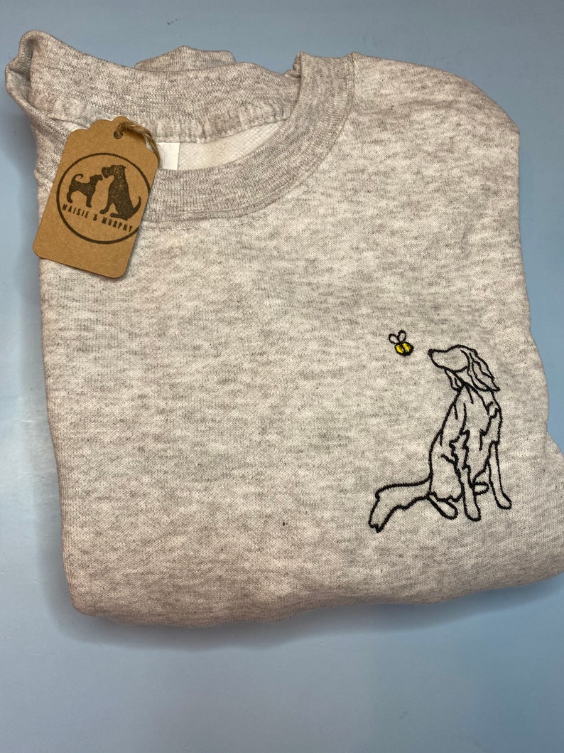 May include: A heather gray sweatshirt with a black embroidered dog and yellow bee design. The dog is looking up at the bee. A brown tag with the text "Maisie & Murphy" is attached to the sweatshirt.
