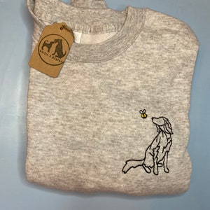 May include: A light gray sweatshirt with a crew neck and a folded design. The sweatshirt features an embroidered black outline of a dog looking at a yellow and black bee. A brown tag with the text "Waisie & Murphy" hangs from the sweatshirt.