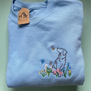 May include: A light blue sweatshirt with an embroidered image of a dog standing in a field of flowers. The dog is brown and white, and the flowers are pink, yellow, and blue. The sweatshirt has a small brown tag with the text "Walks & Woofs" on it.
