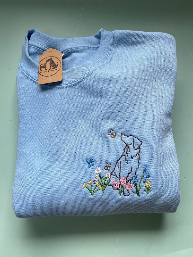 May include: A light blue sweatshirt with an embroidered image of a dog standing in a field of flowers. The dog is brown and white, and the flowers are pink, yellow, and blue. The sweatshirt has a small brown tag with the text "Walks & Woofs" on it.
