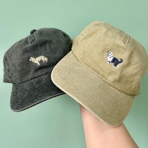 May include: Two baseball caps, one dark grey and one khaki, are held up against a light green background. Each cap features an embroidered dog design. The dark grey cap has a light brown dog, while the khaki cap has a black and white dog.