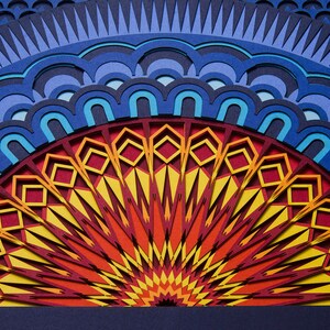 Op de afbeelding: Een meerlaags papieren kunstwerk met een halfrond mandala-ontwerp. Het midden straalt met levendige gele, oranje en rode geometrische patronen, omringd door concentrische ringen van blauwe, lichtblauwe en donkerblauwe geschulpte vormen.