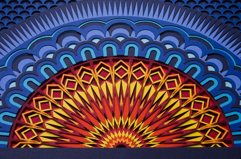 Op de afbeelding: Een meerlaags papieren kunstwerk met een halfrond mandala-ontwerp. Het midden straalt met levendige gele, oranje en rode geometrische patronen, omringd door concentrische ringen van blauwe, lichtblauwe en donkerblauwe geschulpte vormen.