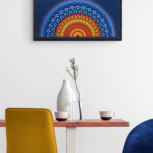 Op de afbeelding: Een rechthoekig wandkunstwerk met een levendig, meerlaags mandala-ontwerp in rood-, oranje-, geel- en blauwtinten, ingelijst in zwart. Daaronder staat een houten tafel met een witte vaas, een glazen vaas met een takje en twee mokken.