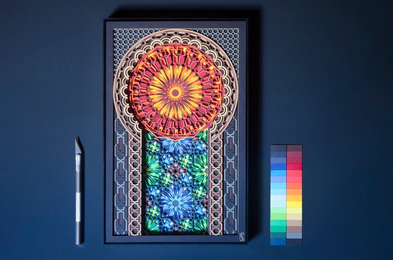 May include: A framed paper art piece featuring a colourful, intricate design. The design includes a large, circular mandala with red, orange, and yellow colours, surrounded by smaller, geometric patterns in shades of blue, green, and brown.