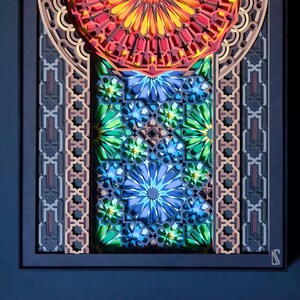 May include: A framed paper art piece with a colourful geometric design. The design features a central circle with radiating lines, surrounded by intricate patterns in shades of red, orange, yellow, green, blue, and brown.